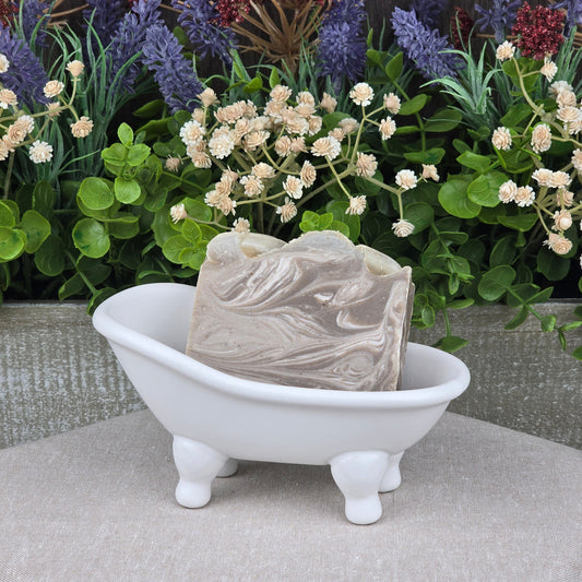 The Jarhead Aromatics Porter + Coffee Handmade Craft Beer Artisan Soap with coffee exfoliant and porter beer sits in a white bathtub-shaped dish, with artificial green leaves and colorful flowers on a wooden background.