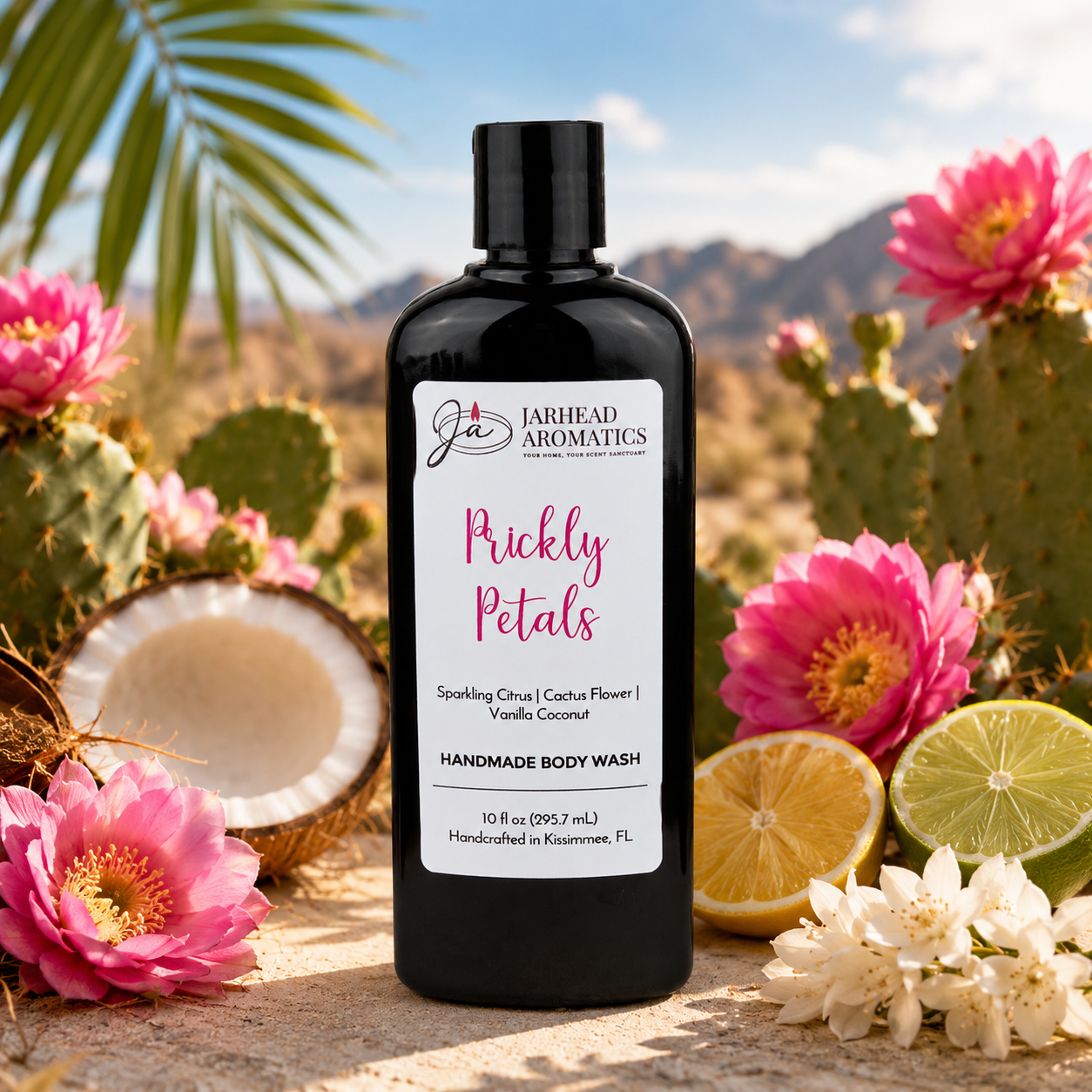A black bottle of Jarhead Aromatics Prickly Petals Handmade Body Wash with organic aloe and plant-based oils sits outdoors amid pink cacti flowers, a halved coconut, white blossoms, and a desert landscape with mountains in the background.