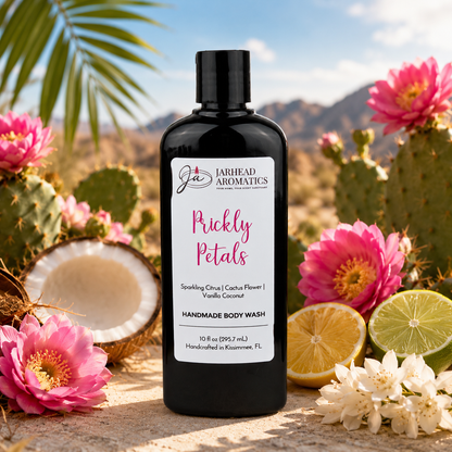 A black bottle of Jarhead Aromatics Prickly Petals Handmade Body Wash with organic aloe and plant-based oils sits outdoors amid pink cacti flowers, a halved coconut, white blossoms, and a desert landscape with mountains in the background.