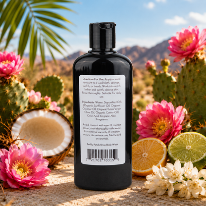 A black bottle of Jarhead Aromatics’ Prickly Petals Handmade Body Wash with organic aloe and plant-based oils sits outdoors among pink cacti flowers, citrus fruits, and palm leaves, its white label with ingredients and directions highlighted in bright sunlight.