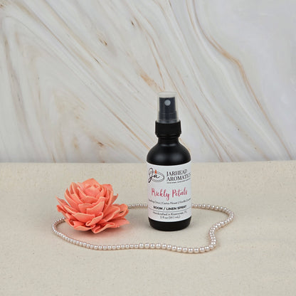 A bottle of Jarhead Aromatics Prickly Petals Room & Linen Spray | Cactus Flower & Sandalwood stands on a beige surface with an artificial coral flower and pearls, set against a marble-patterned background.