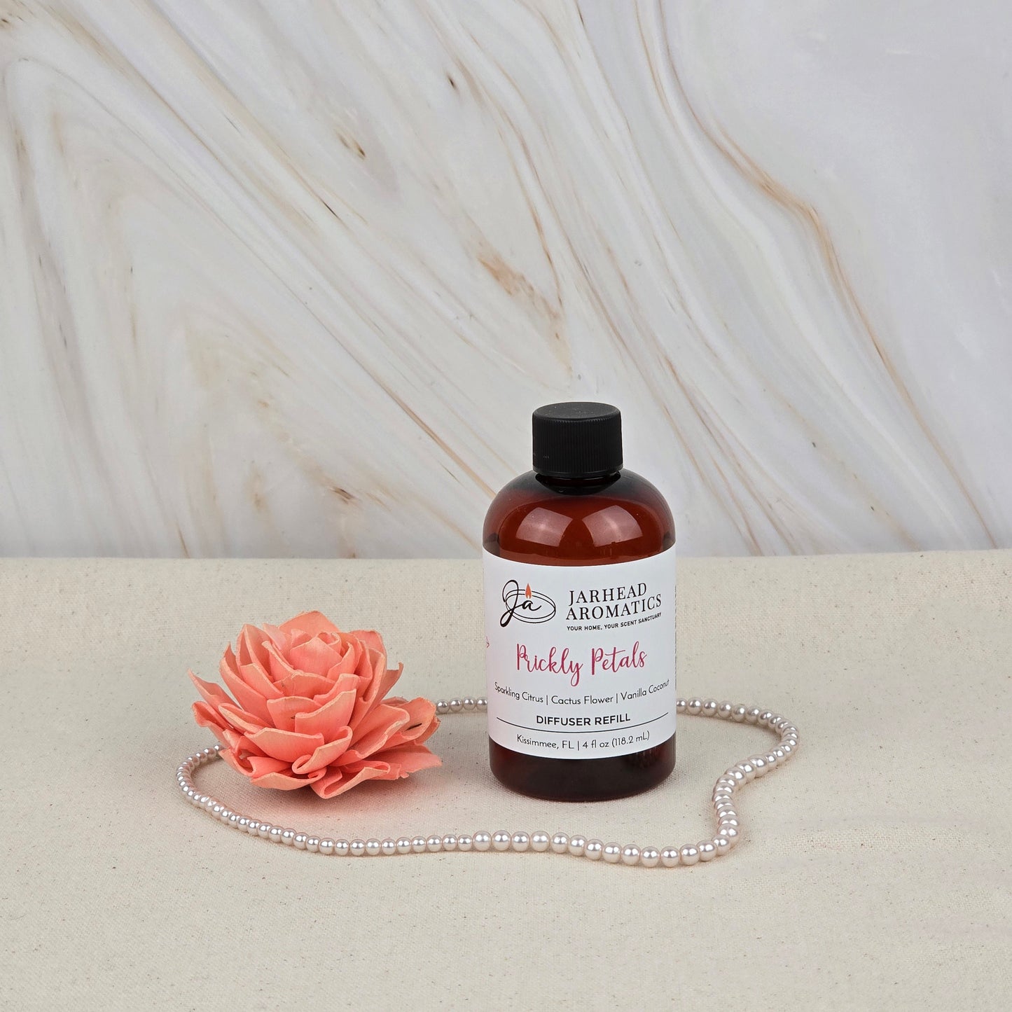 A bottle of Jarhead Aromatics’ Prickly Petals Diffuser Refill | Cactus Flower, Citrus & Sandalwood sits on a beige surface with a pink artificial flower and pearls, set against a white marble background.