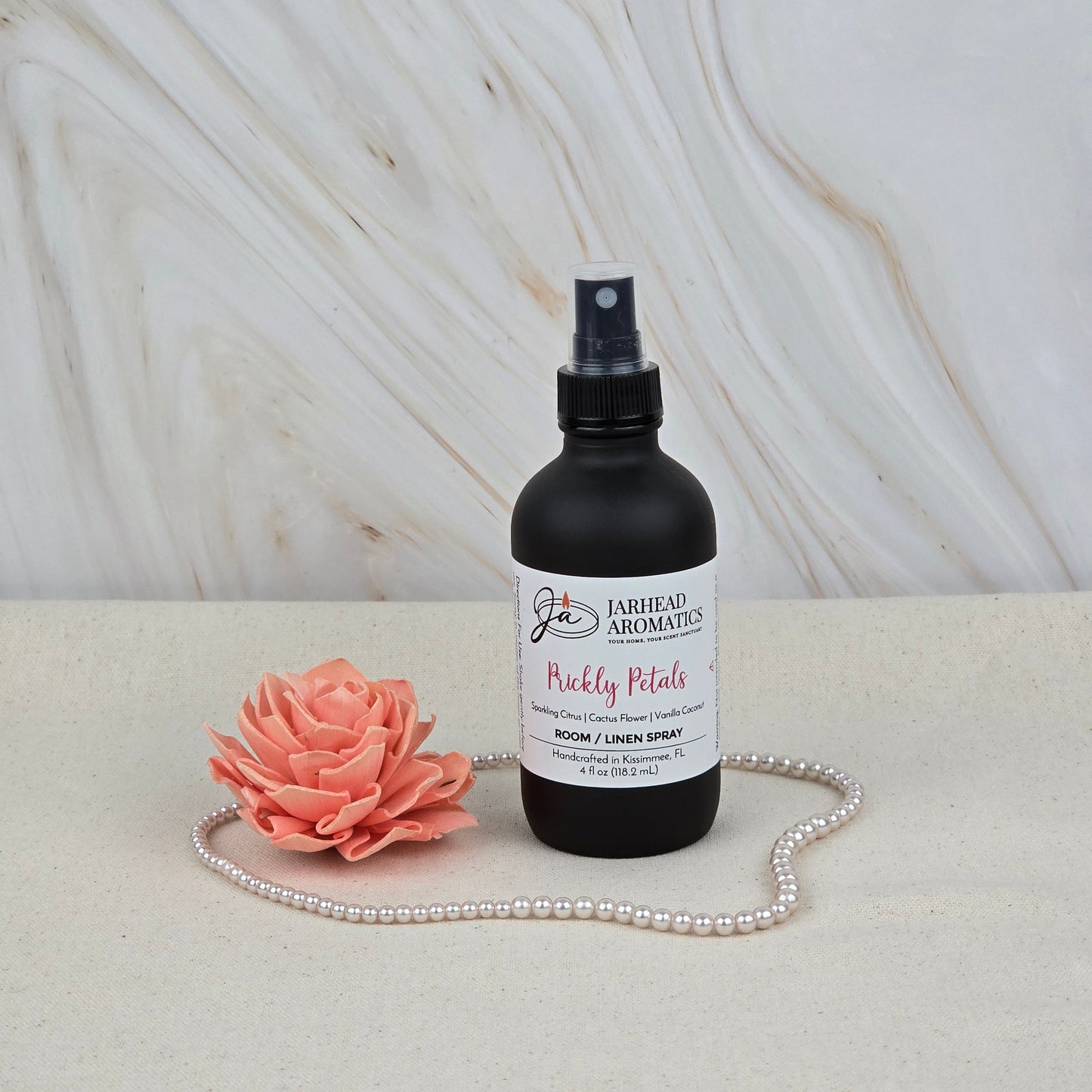 A black Jarhead Aromatics Prickly Petals Room & Linen Spray (Cactus Flower & Sandalwood) sits beside a pink artificial flower and pearls on a light surface, with a marble background—ideal for adding desert-inspired fragrance to your home.
