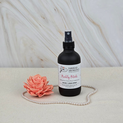 A black Jarhead Aromatics Prickly Petals Room & Linen Spray (Cactus Flower & Sandalwood) sits beside a pink artificial flower and pearls on a light surface, with a marble background—ideal for adding desert-inspired fragrance to your home.