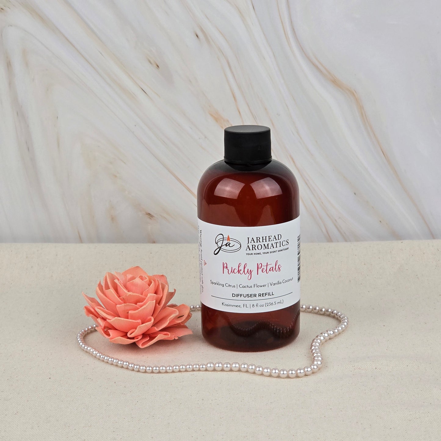 A brown bottle of Jarhead Aromatics Prickly Petals Diffuser Refill | Cactus Flower, Citrus & Sandalwood sits on a beige surface beside a pink faux flower and pearls, adding a desert-inspired scent to your home fragrance collection.