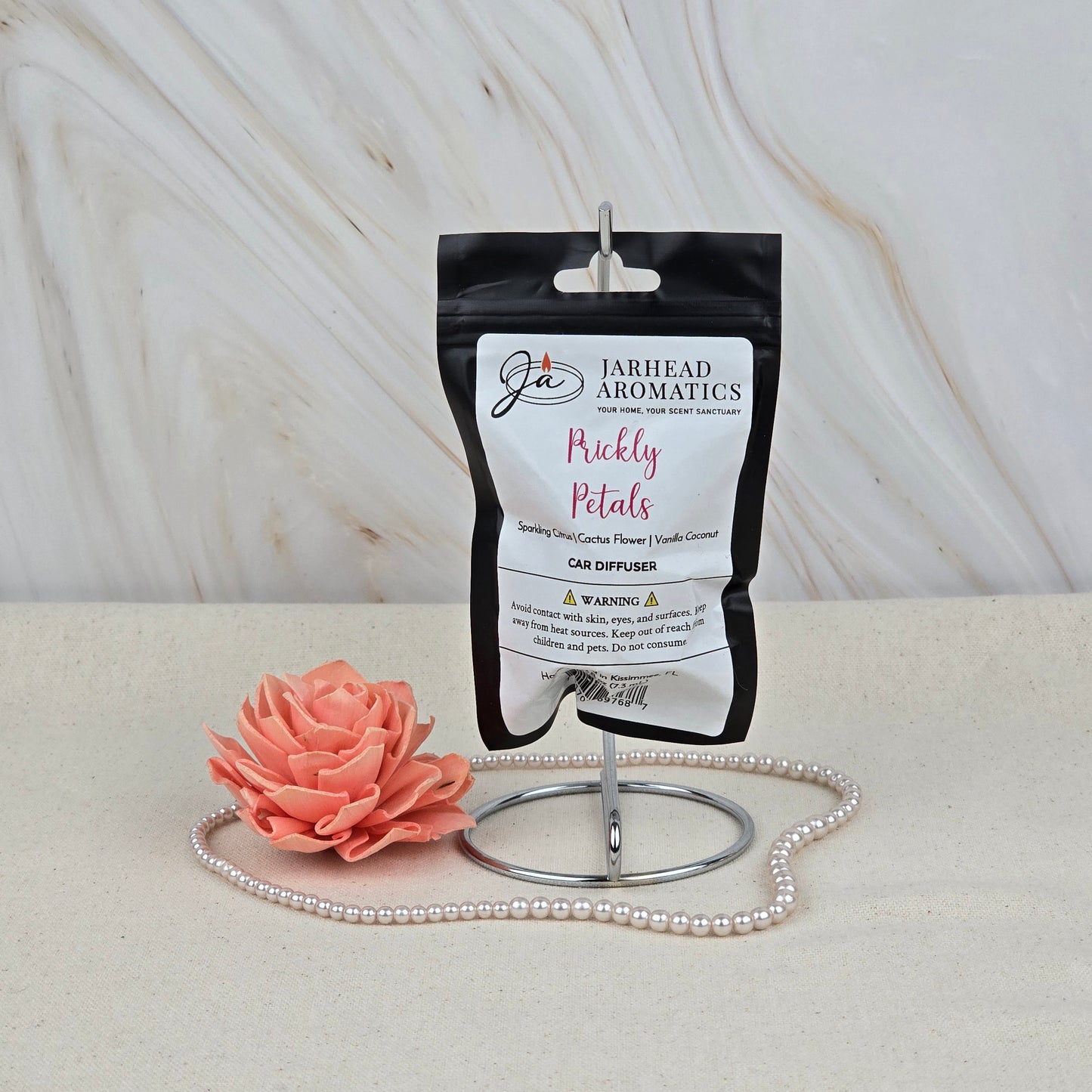 A Jarhead Aromatics Prickly Petals Car Diffuser, a cactus flower & citrus air freshener, is showcased on a metal stand with a pink flower and pearls against a marble backdrop.