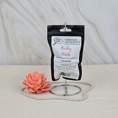 A Jarhead Aromatics Prickly Petals Car Diffuser, a cactus flower & citrus air freshener, is showcased on a metal stand with a pink flower and pearls against a marble backdrop.