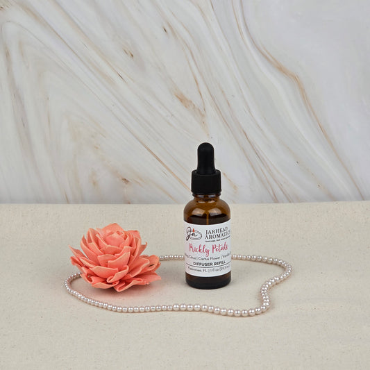 A Prickly Petals Car Diffuser Refill by Jarhead Aromatics rests on a light surface beside a pink flower and pearls, suggesting its cactus flower & citrus scent, with a marbled background setting the scene.