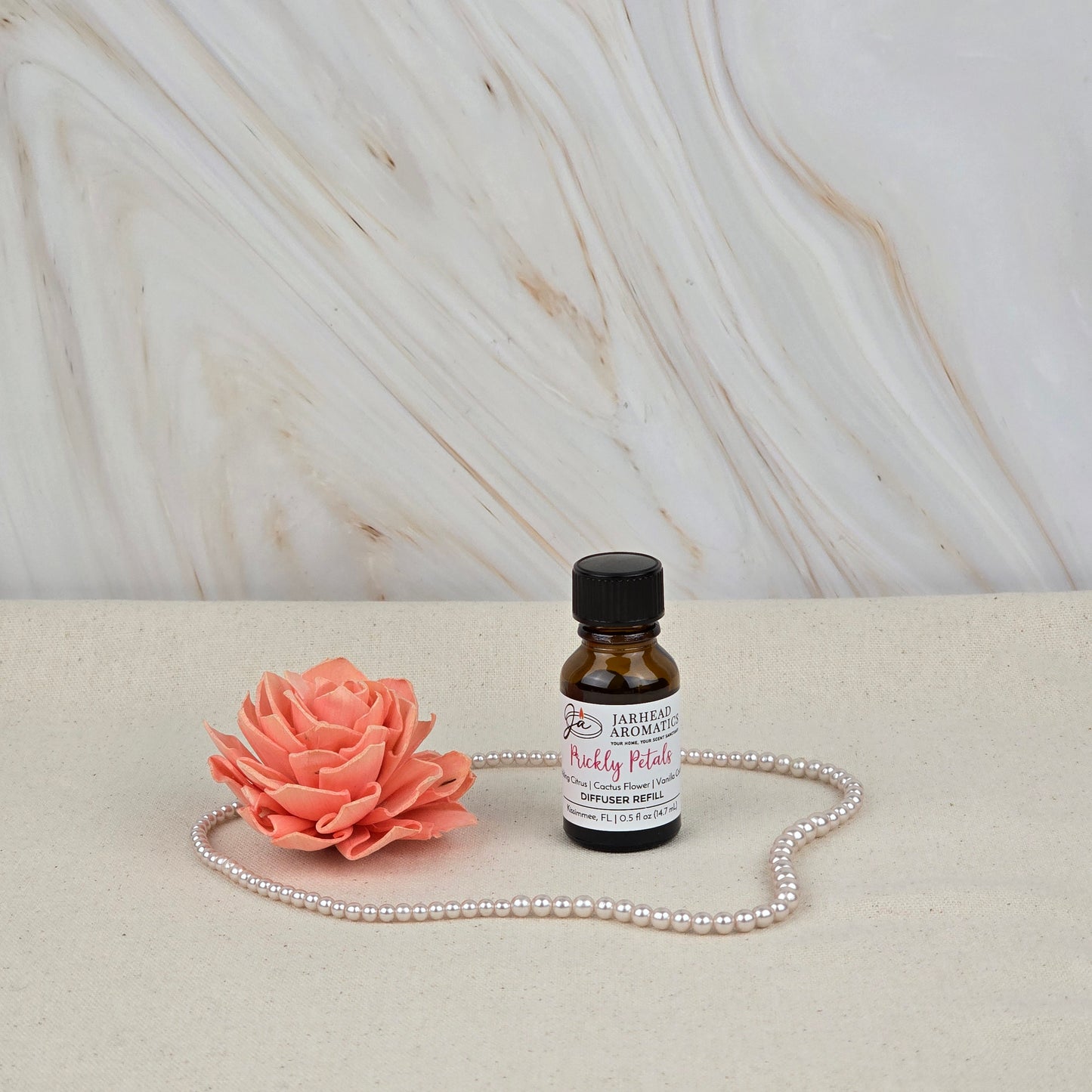 A small amber bottle of Jarhead Aromatics Prickly Petals Cold Air Diffuser Refill | Cactus Flower & Coconut sits on a light surface with a pink flower and pearls, bringing long-lasting luxury fragrance against a marble-patterned background.
