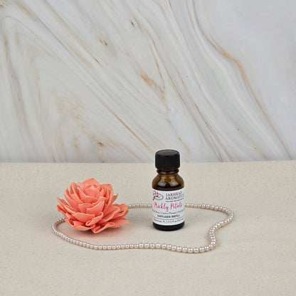 A small amber bottle of Jarhead Aromatics Prickly Petals Cold Air Diffuser Refill | Cactus Flower & Coconut sits on a light surface with a pink flower and pearls, bringing long-lasting luxury fragrance against a marble-patterned background.