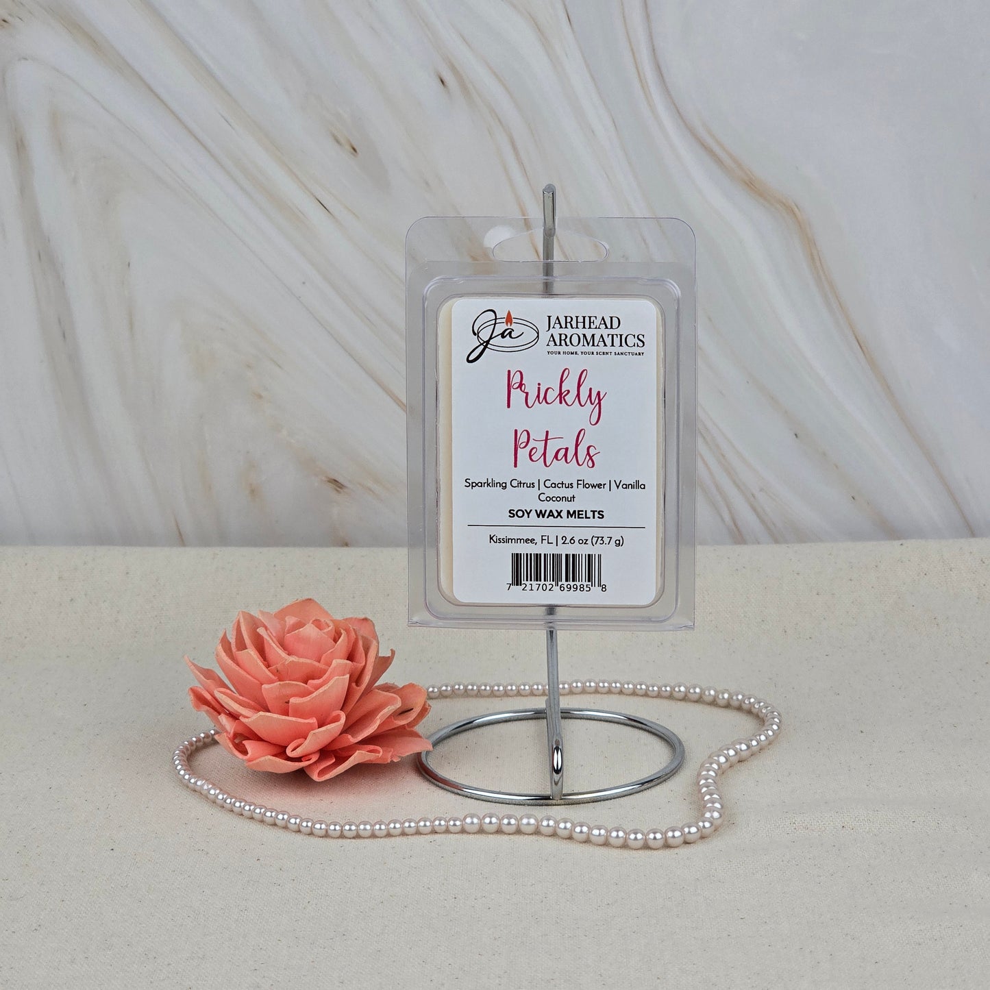 A pack of Jarhead Aromatics Prickly Petals Soy Wax Melts (Cactus Flower & Coconut) sits on a metal stand beside a peach artificial flower and pink beaded necklace, set against a marble-textured background.