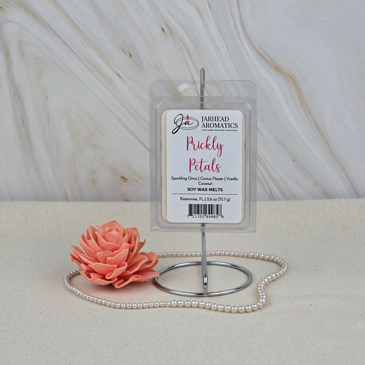 A pack of Jarhead Aromatics Prickly Petals Soy Wax Melts (Cactus Flower & Coconut) sits on a metal stand beside a peach artificial flower and pink beaded necklace, set against a marble-textured background.