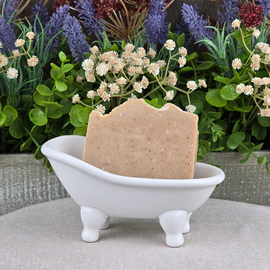 A bar of Pumpkin + Spice Handmade Seasonal Artisan Soap by Jarhead Aromatics rests in a white clawfoot bathtub soap dish, with green foliage and purple and cream flowers in the background.