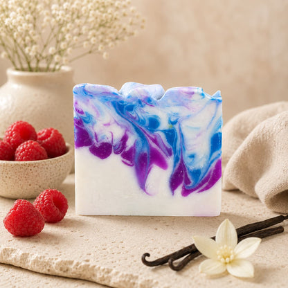 A bar of Jarhead Aromatics Raspberry + Vanilla Handmade Coconut Milk Artisan Soap, featuring a blue, purple, and white swirl pattern, rests on a stone surface surrounded by fresh raspberries, vanilla beans, a white flower, a beige vase filled with baby’s breath, and a soft towel in the background.