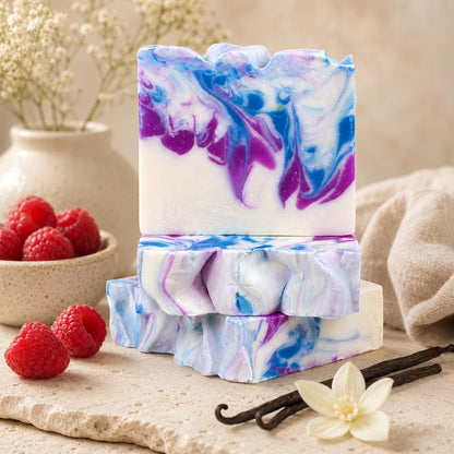 Three vibrant Raspberry + Vanilla Handmade Coconut Milk Artisan Soap bars from Jarhead Aromatics, featuring blue and purple marbled swirls, are stacked on a stone surface alongside fresh raspberries, a beige vase with white flowers, a vanilla bean, and a cream-colored cloth. Handcrafted with nourishing coconut milk, this luxurious artisan soap offers an indulgent touch.