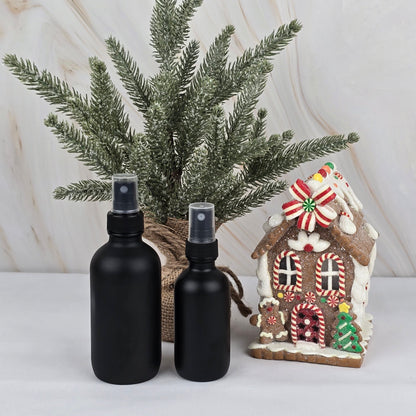 Two black bottles of Jarhead Aromatics Merry & Bright Room/Linen Spray from the Winter Collection are set before a small pine tree in burlap, next to a gingerbread house, making a festive holiday scene against a light background.