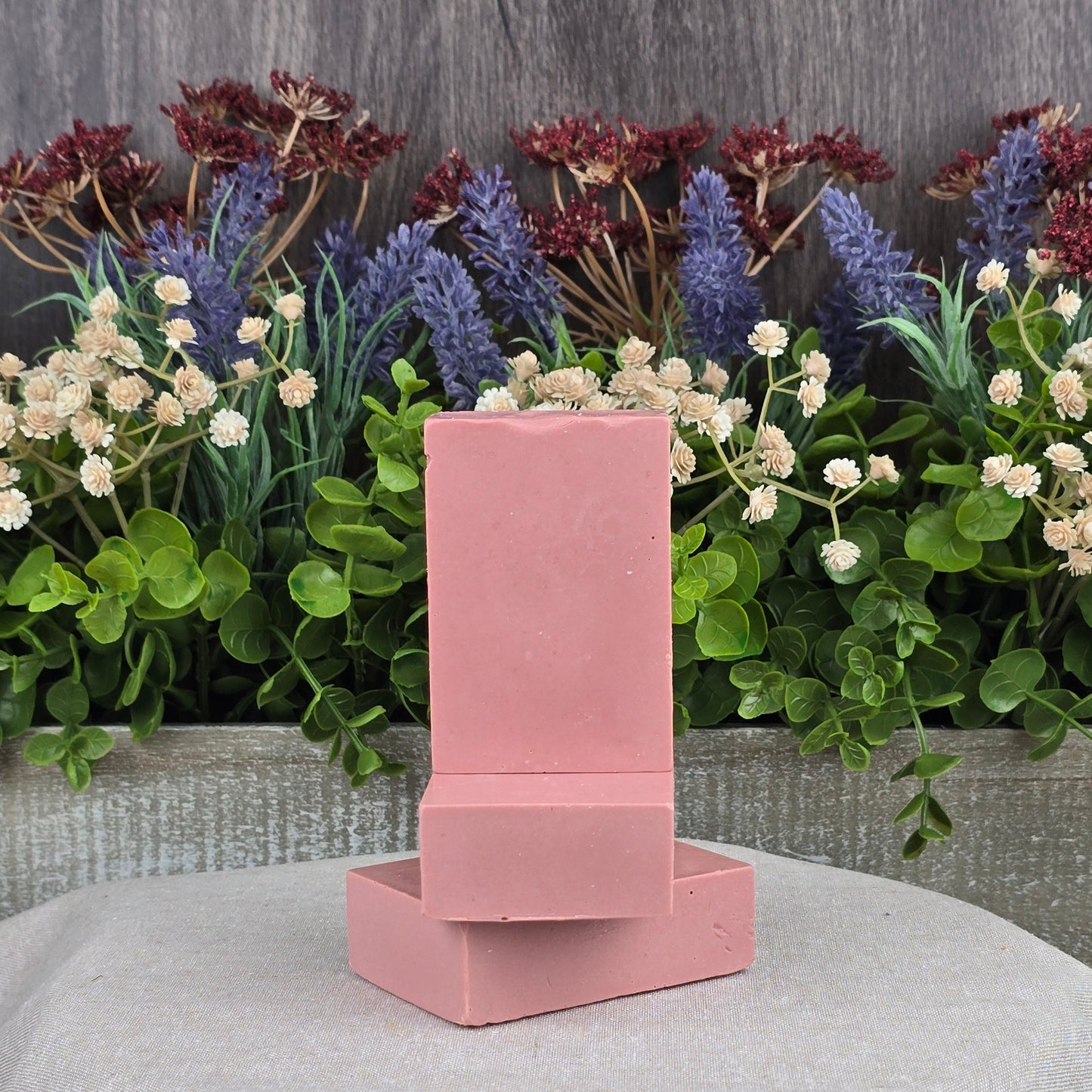 Three Rose + Calamine Handmade Goat Milk Facial Soaps from Jarhead Aromatics are showcased with one standing and one lying down. They are surrounded by lush greenery and clusters of purple and red flowers on a gray wooden panel atop a gray fabric surface.