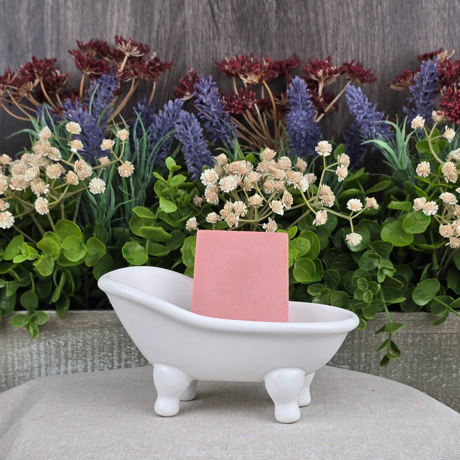 A small white, clawfoot bathtub-shaped dish holds a pink Rose + Calamine Handmade Goat Milk Facial Soap by Jarhead Aromatics. Behind it is a display of assorted artificial flowers and greenery against a wooden background on a textured cloth surface, highlighting the elegance of nourishing oils.