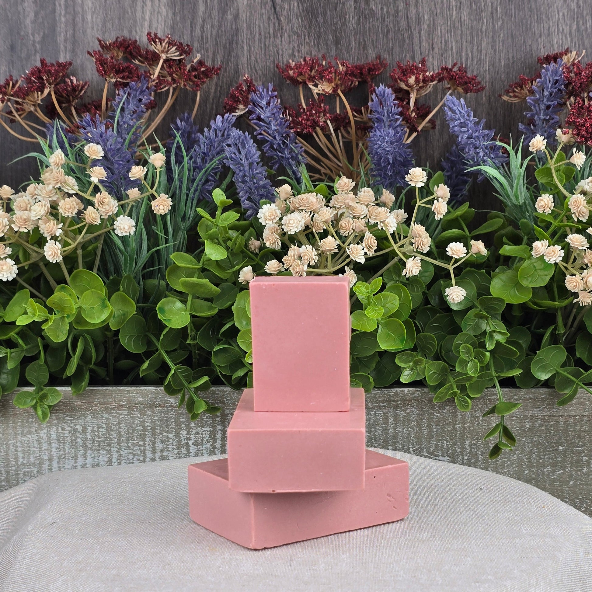 Three Jarhead Aromatics Rose + Calamine Handmade Goat Milk Facial Soaps, featuring nourishing oils and botanicals, are stacked against lush foliage with purple and white flowers. The setup on a gray cloth exudes a fresh, natural ambiance.