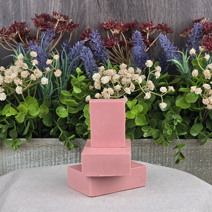 Three Jarhead Aromatics Rose + Calamine Handmade Goat Milk Facial Soaps, featuring nourishing oils and botanicals, are stacked against lush foliage with purple and white flowers. The setup on a gray cloth exudes a fresh, natural ambiance.