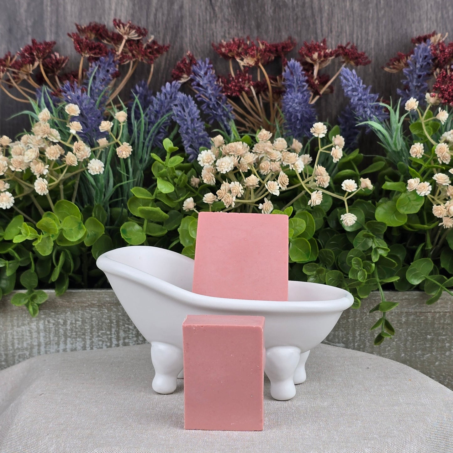 Two Rose + Calamine Handmade Goat Milk Facial Soaps by Jarhead Aromatics, made with natural ingredients, are placed near a small white clawfoot tub. The backdrop features purple, red, and white flowers among lush green foliage on a textured gray cloth.