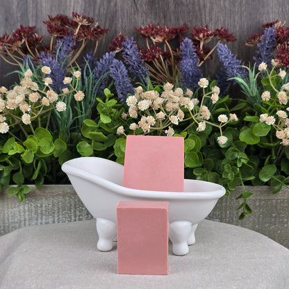 Two Rose + Calamine Handmade Goat Milk Facial Soaps by Jarhead Aromatics, made with natural ingredients, are placed near a small white clawfoot tub. The backdrop features purple, red, and white flowers among lush green foliage on a textured gray cloth.