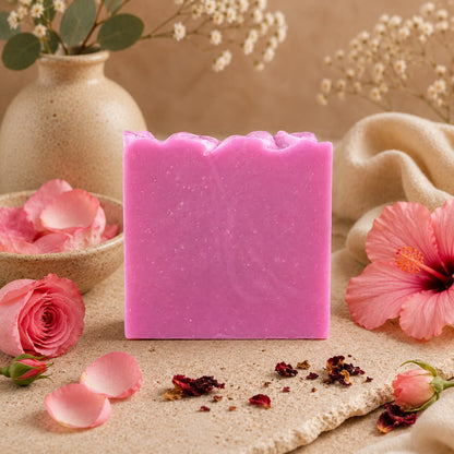 A bar of Jarhead Aromatics’ Rose + Hibiscus Handmade Coconut Milk Artisan Soap stands upright on a textured surface, surrounded by rose petals, dried flowers, and beige vases filled with blooms, creating a soft and elegant atmosphere that highlights its romantic rose and tropical floral scent.