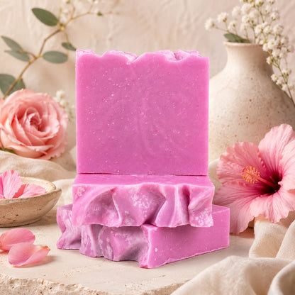 Two Rose + Hibiscus Handmade Coconut Milk Artisan Soap bars from Jarhead Aromatics are stacked on a neutral surface, complemented by fresh flowers and ceramic vases. The scene exudes a soft, elegant aesthetic with floral accents and gentle lighting, highlighting the romantic rose and tropical floral notes of the soap.