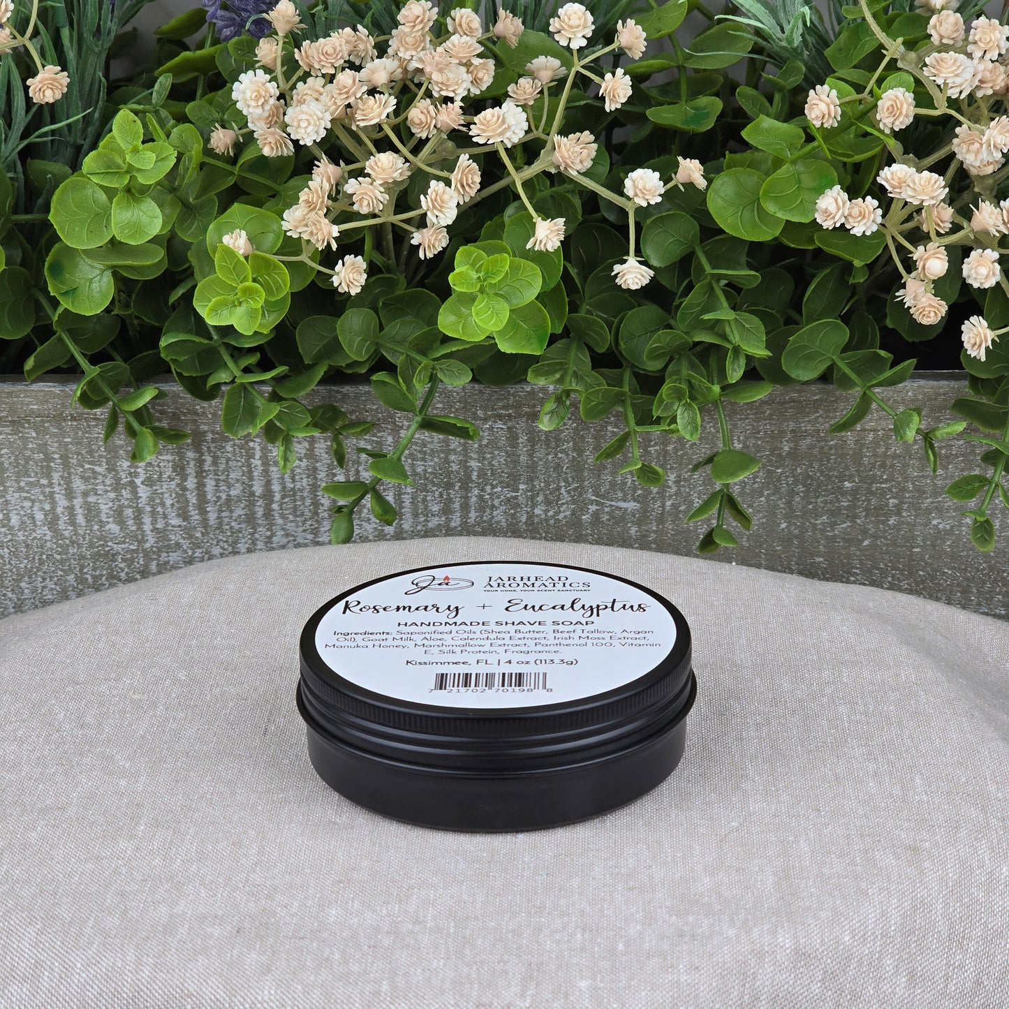 A black tin of Jarhead Aromatics’ Rosemary + Eucalyptus Handmade Shave Soap | Natural Tallow & Goat Milk Shaving Soap sits on a beige surface, beside green leaves and cream flowers, reflecting the shaving soap’s natural inspiration.