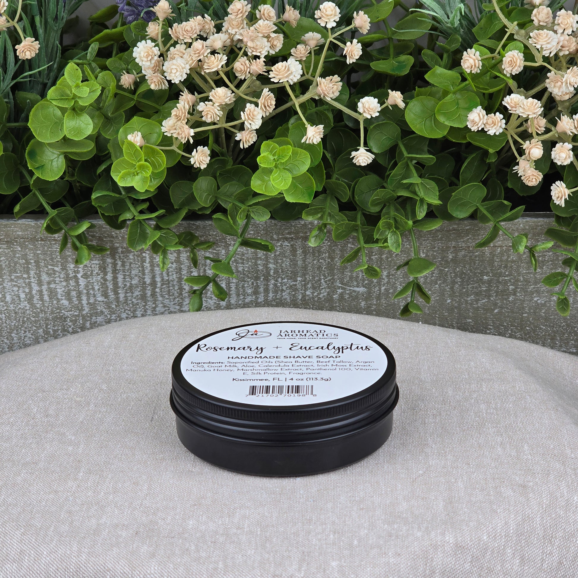 A black tin of Jarhead Aromatics’ Rosemary + Eucalyptus Handmade Shave Soap | Natural Tallow & Goat Milk Shaving Soap sits on a beige surface, beside green leaves and cream flowers, reflecting the shaving soap’s natural inspiration.