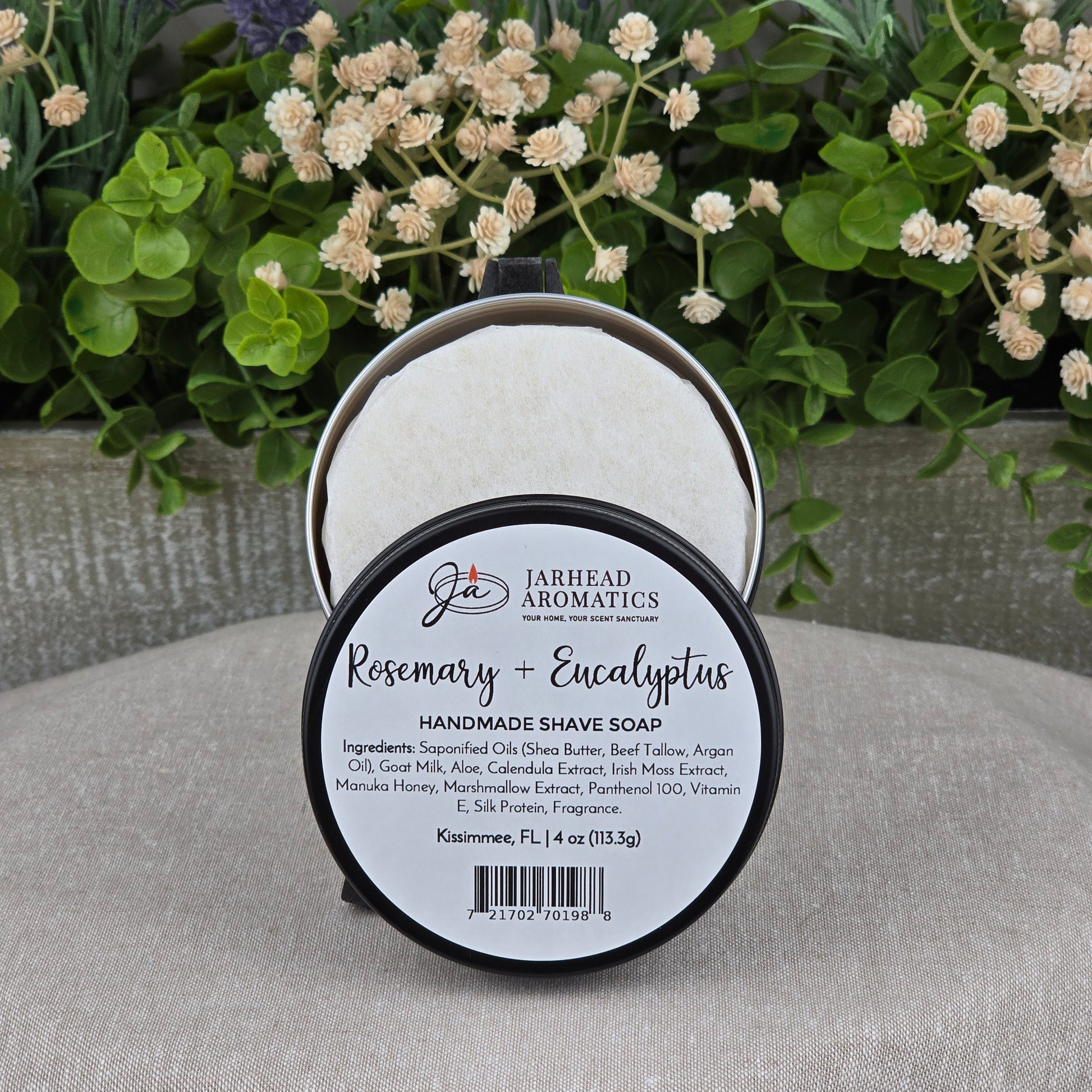 A round Jarhead Aromatics Rosemary + Eucalyptus Handmade Shave Soap with natural tallow and goat milk rests on stone, its label detailing ingredients against a blurred background of green leaves and purple flowers.