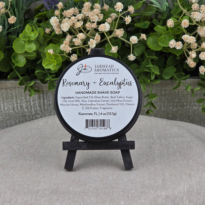 A round tin of Rosemary + Eucalyptus Handmade Shave Soap by Jarhead Aromatics, made with natural tallow and goat milk, rests on a small black easel amid green foliage and flowers. Ingredients and product details are listed on the label.