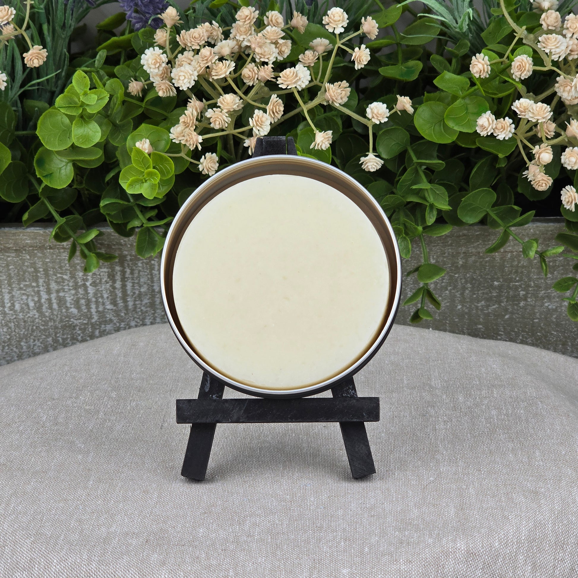 A round, cream Rosemary + Eucalyptus Handmade Shave Soap by Jarhead Aromatics sits on a black easel before green foliage and white flowers, displayed on a textured beige surface. Made with natural tallow and goat milk.
