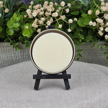 A round, cream Rosemary + Eucalyptus Handmade Shave Soap by Jarhead Aromatics sits on a black easel before green foliage and white flowers, displayed on a textured beige surface. Made with natural tallow and goat milk.