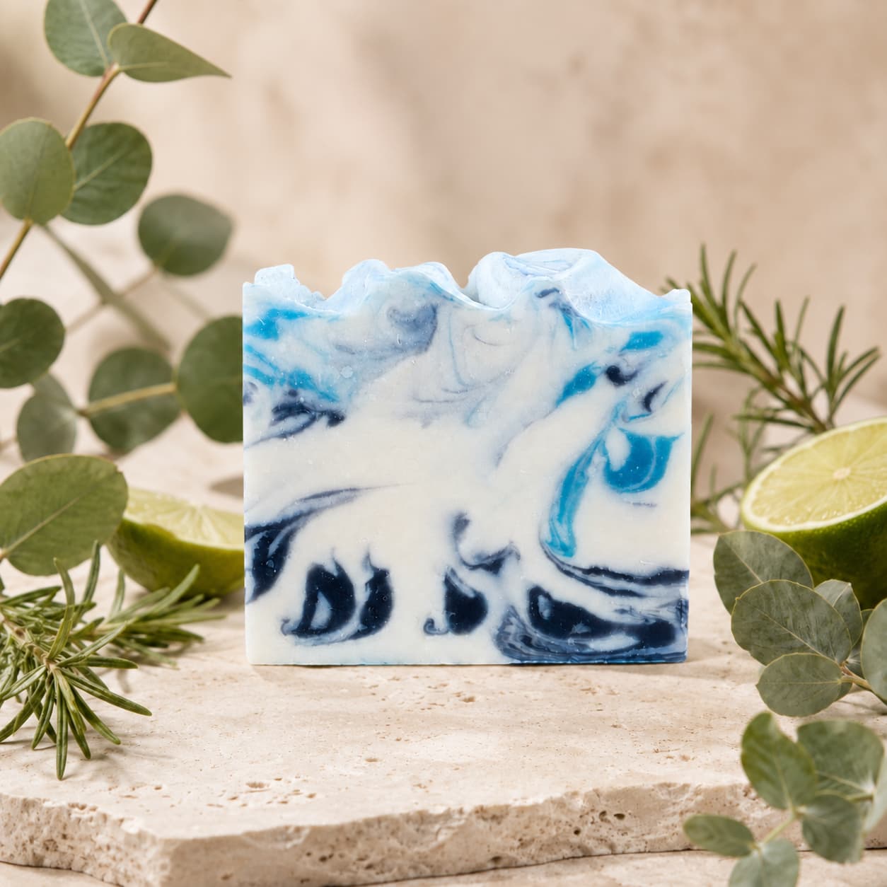 A bar of Jarhead Aromatics’ Rosemary + Eucalyptus Handmade Coconut Milk Artisan Soap rests on a stone surface, accented by fresh eucalyptus leaves, sprigs of rosemary, and halved limes against a soft beige background. This fresh herbal spa soap is beautifully handcrafted for a luxurious bathing experience.