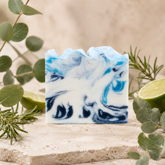 A bar of Jarhead Aromatics’ Rosemary + Eucalyptus Handmade Coconut Milk Artisan Soap rests on a stone surface, accented by fresh eucalyptus leaves, sprigs of rosemary, and halved limes against a soft beige background. This fresh herbal spa soap is beautifully handcrafted for a luxurious bathing experience.