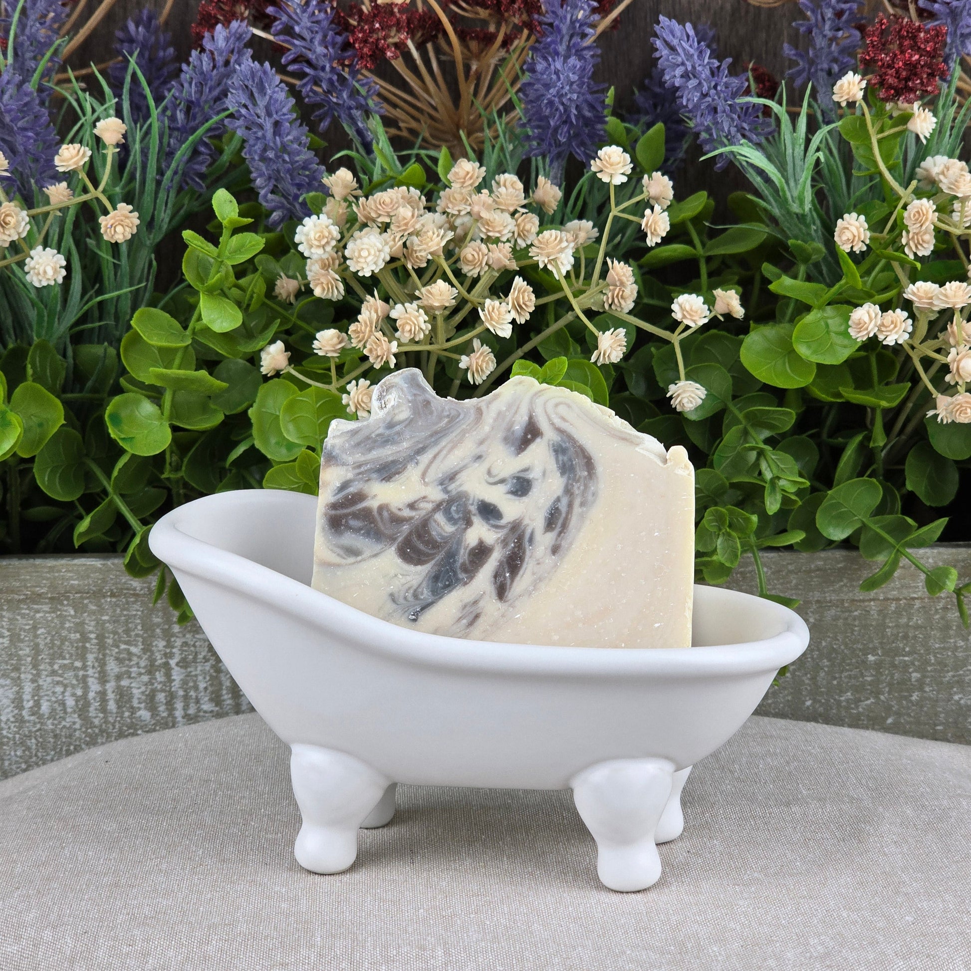 The Jarhead Aromatics Sandalwood + Patchouli Handmade Artisan Soap rests in a small white clawfoot bathtub dish, set before lush green plants and purple flowers.