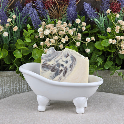 The Jarhead Aromatics Sandalwood + Patchouli Handmade Artisan Soap rests in a small white clawfoot bathtub dish, set before lush green plants and purple flowers.