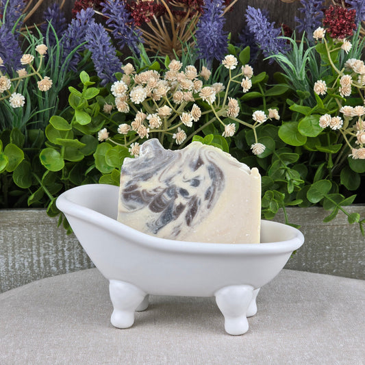 The Jarhead Aromatics Sandalwood + Patchouli Handmade Artisan Soap rests in a small white clawfoot bathtub dish, set before lush green plants and purple flowers.