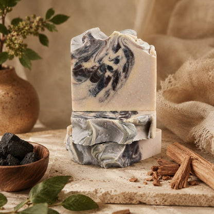 Two bars of Jarhead Aromatics Sandalwood + Patchouli Handmade Artisan Soap, featuring marbled black, white, and gray swirls, are stacked on a stone surface amid charcoal pieces, cinnamon sticks, fresh green leaves, and a rustic vase in the background. This earthy woodsy charcoal soap is crafted with activated charcoal for deep cleansing and offers a luxurious bathing experience with its rich sandalwood patchouli scent.