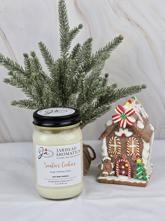 A Santas Cookies soy wax candle from Jarhead Aromatics, infused with essential oils, sits beside a Christmas tree and gingerbread house, evoking holiday cheer.
