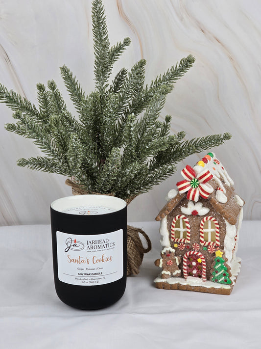 The Santas Cookies soy wax candle by Jarhead Aromatics fills the air with holiday scents, complemented by a gingerbread house and a frosted evergreen in the background, for a cozy festive ambiance.