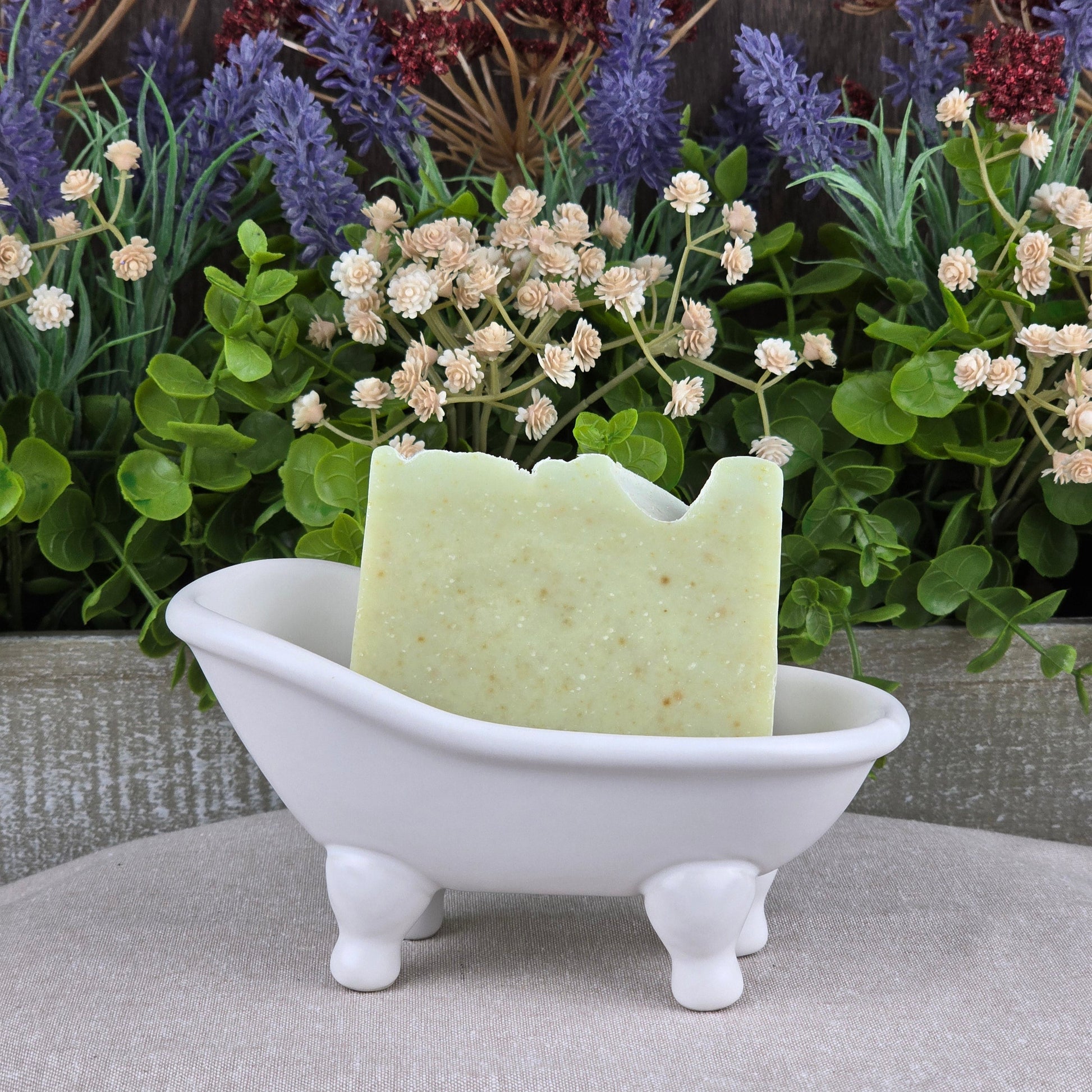 A bar of Jarhead Aromatics' Sea Kelp + Aloe Handmade Artisan Soap sits in a small white bathtub-shaped dish, with artificial greenery and purple and white flowers in the background.