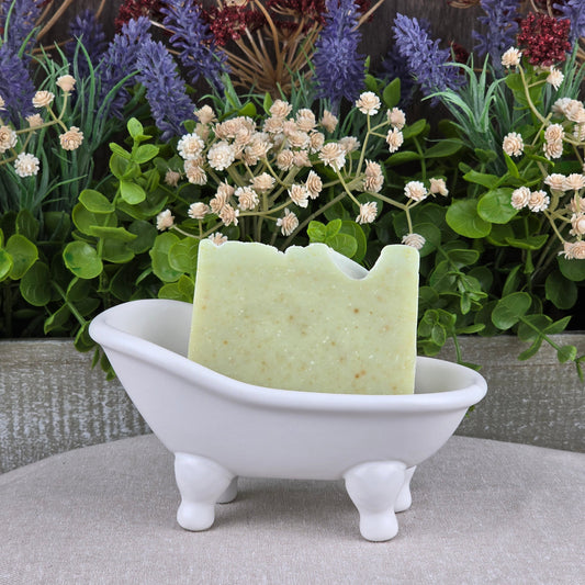 A bar of Jarhead Aromatics' Sea Kelp + Aloe Handmade Artisan Soap sits in a small white bathtub-shaped dish, with artificial greenery and purple and white flowers in the background.