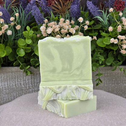 Three bars of Jarhead Aromatics Sea Kelp + Aloe Handmade Natural Goat Milk Artisan Soap are stacked in front of a wooden planter filled with colorful artificial flowers and greenery, set against an outdoor natural background.