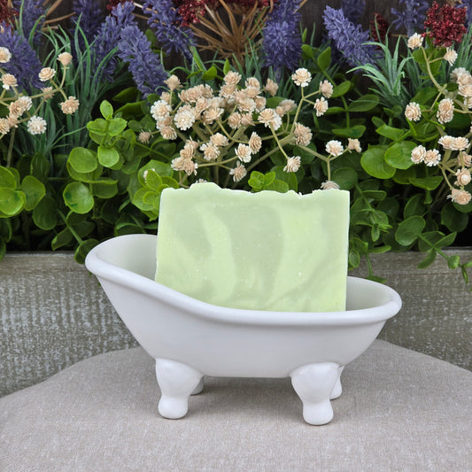 A Jarhead Aromatics Sea Kelp + Aloe Handmade Natural Goat Milk Artisan Soap bar rests in a small white bathtub-shaped soap dish, with assorted artificial green plants and flowers displayed in the background.