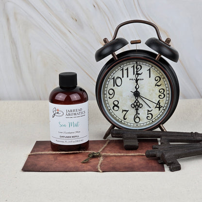 A vintage black alarm clock, two large metal keys, a leather-bound book, and a Jarhead Aromatics Sea Mist Diffuser Refill | Ozone, Sea Salt & Linen enhance a light surface with marble background, evoking fresh coastal fragrance.