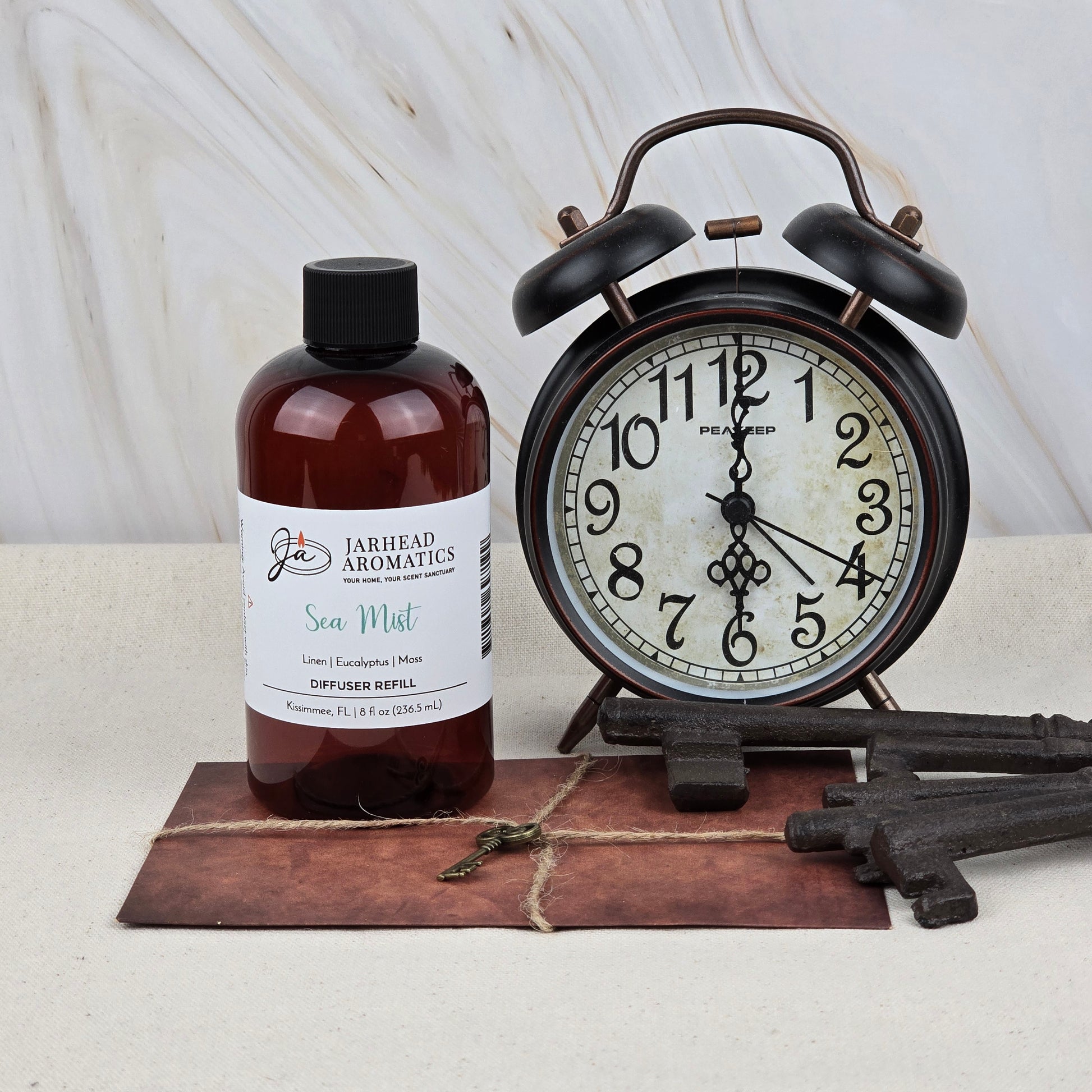 A brown bottle of Jarhead Aromatics Sea Mist Diffuser Refill—Ozone, Sea Salt & Linen Home Fragrance Oil—sits beside a vintage black alarm clock reading 10:11, with old keys and a tied envelope nearby on a light surface.