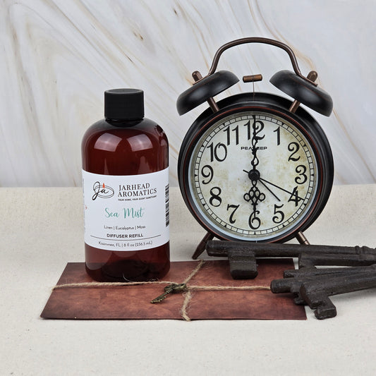 A brown bottle of Jarhead Aromatics Sea Mist Diffuser Refill—Ozone, Sea Salt & Linen Home Fragrance Oil—sits beside a vintage black alarm clock reading 10:11, with old keys and a tied envelope nearby on a light surface.