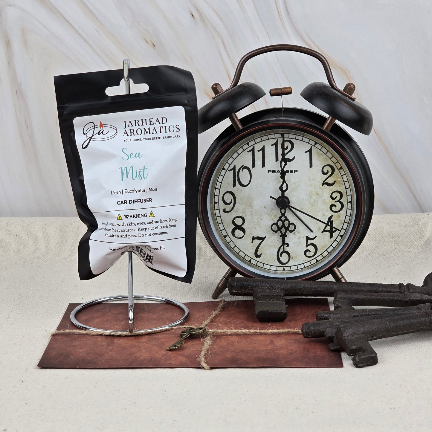A packet of Jarhead Aromatics Sea Mist Car Diffuser | Clean Coastal & Spa-Inspired Car Air Freshener hangs on a metal stand beside a vintage black alarm clock, old keys, and a brown envelope on a neutral surface.